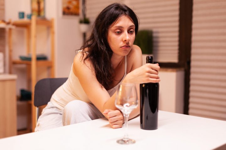 Lonely woman holding a bottle of red wine. Unhappy person suffering of migraine, depression, disease and anxiety feeling exhausted with dizziness symptoms having alcoholism problems.
